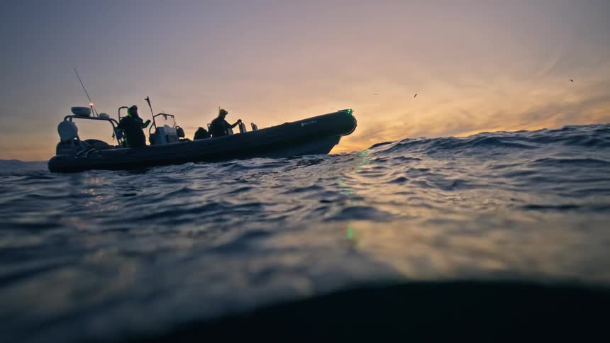 An over-under slow-motion shot captures a rigid inflatable boat with divers floating on the open sea against a beautiful sunset sky