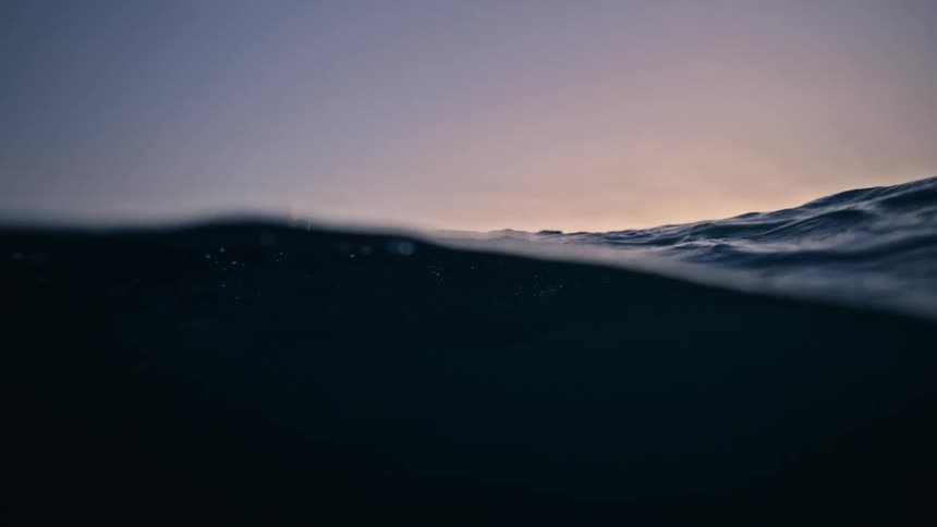 Slow motion, over-under shot reveals a rigid inflatable boat with passengers silhouetted against a dramatic sunset on the open ocean