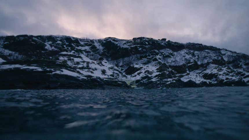 First-person view emerging from the cold, dark ocean to reveal a snowy coastline under a gloomy sky, then sinking back down