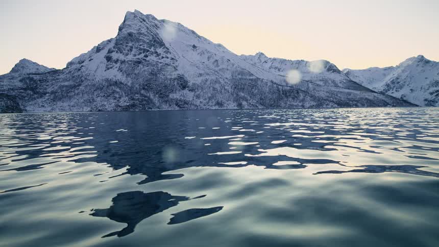 A serene low-angle view of calm waters rippling in front of a majestic snow-covered mountain range at sunrise