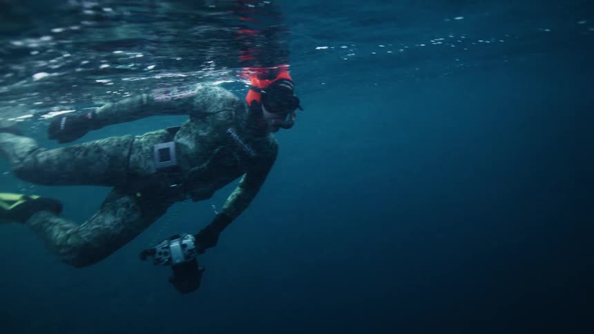 An underwater photographer in a camouflage wetsuit and orange hood swims slowly through the deep blue water with a camera