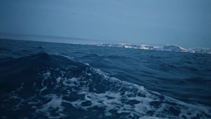 A slow motion view from a boat sailing across the choppy dark blue Arctic Ocean towards a snowy coastline