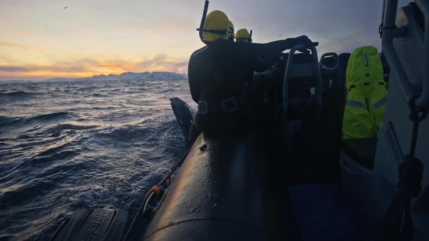 Divers in a rigid inflatable boat navigate the arctic ocean at sunset, watching a whale spout in the distance