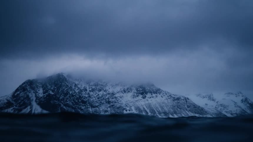 The camera is submerged and resurfaces in the dark, cold ocean, revealing a dramatic snow-covered mountain under a stormy sky