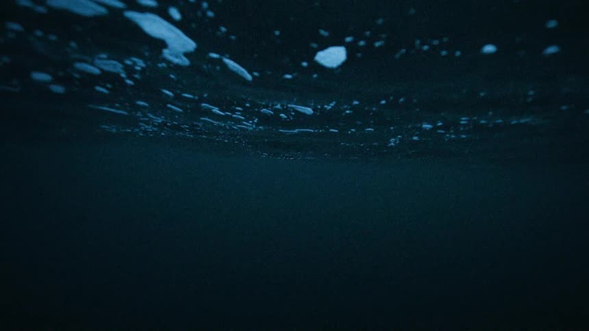 An eerie slow-motion view from beneath the surface of dark, deep blue ocean water