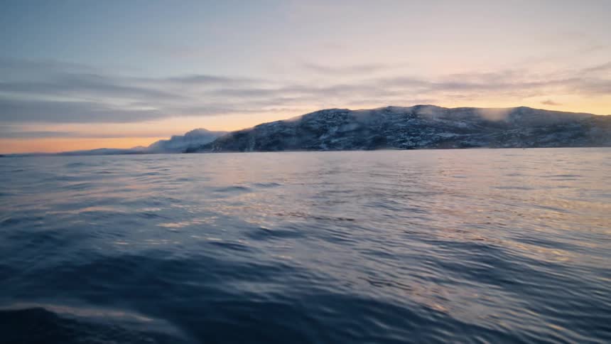 A serene low-angle shot from a boat moving across a steaming arctic ocean with snowy mountains in the distance at sunset