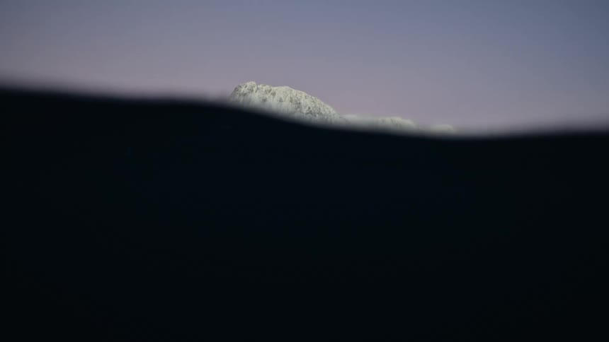 Calm, dark waters gently ripple in slow motion, offering a serene split view of a majestic snow-covered mountain at dusk