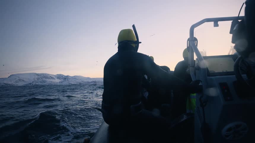 Silhouetted snorkelers ride in a boat across a cold ocean towards snow-covered mountains at sunrise in slow motion