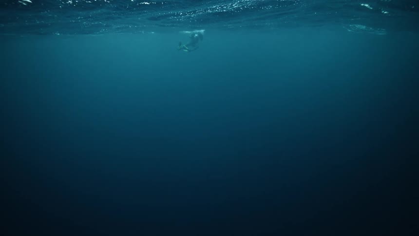 A lone scuba diver floats on the surface of the vast deep blue ocean, as viewed from below