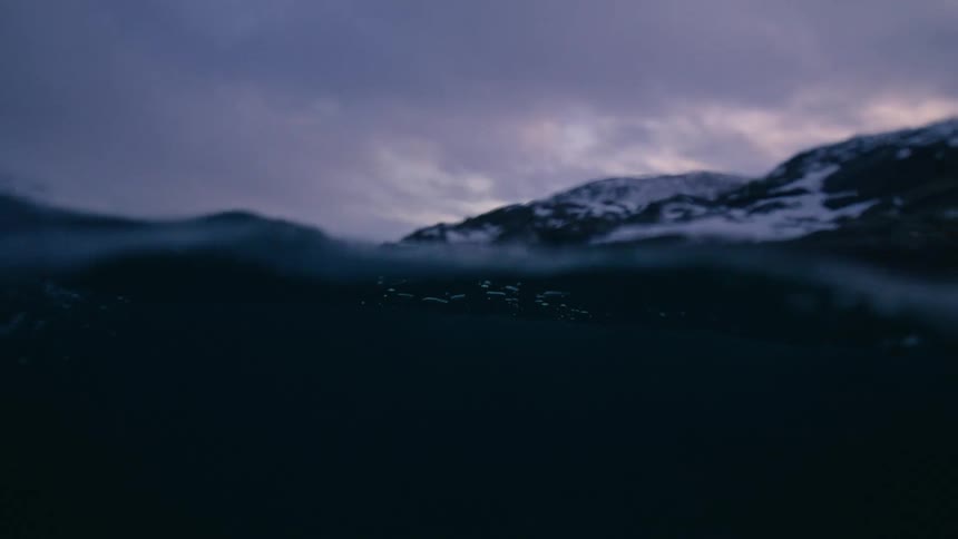 Slow motion shot at water level of a cold, dark fjord with snowy mountains under a moody, overcast sky