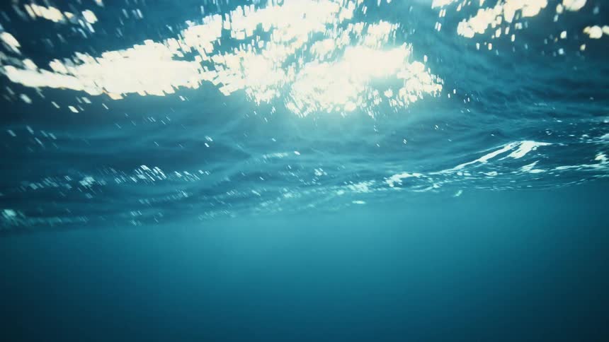 Glistening sunlight reflects off the rippling surface of the deep blue ocean, viewed from underwater