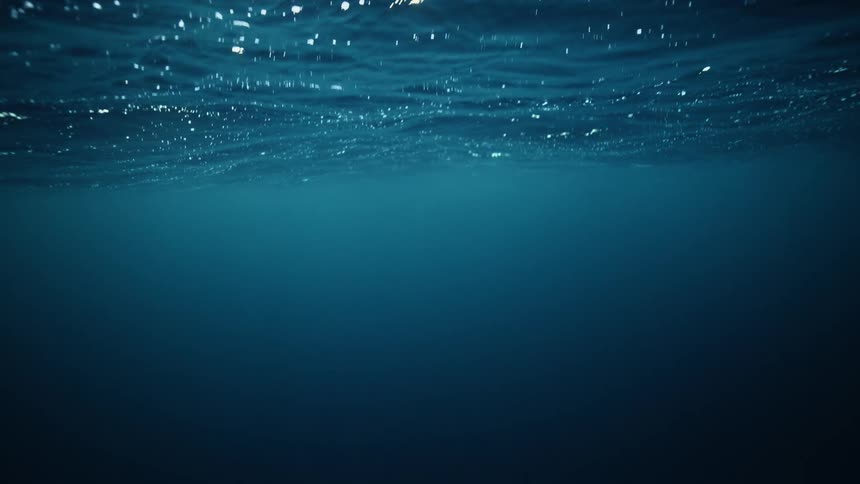 An underwater perspective looking up at the calm, dark blue ocean surface as sunlight shines through