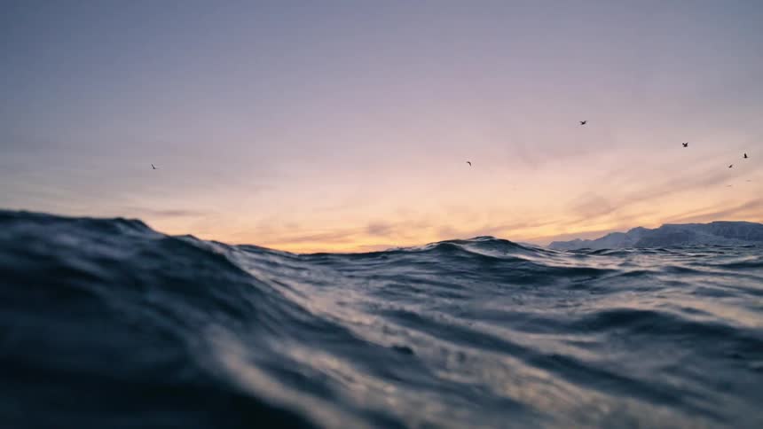 A stunning slow-motion, water-level view of the ocean reveals rolling waves and a beautiful sunset over a distant, snowy mountain range