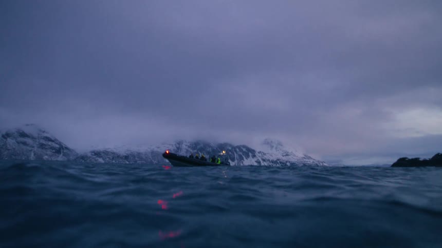A dramatic split-level view from the dark, cold ocean reveals a tour boat floating near snow-covered mountains at dusk