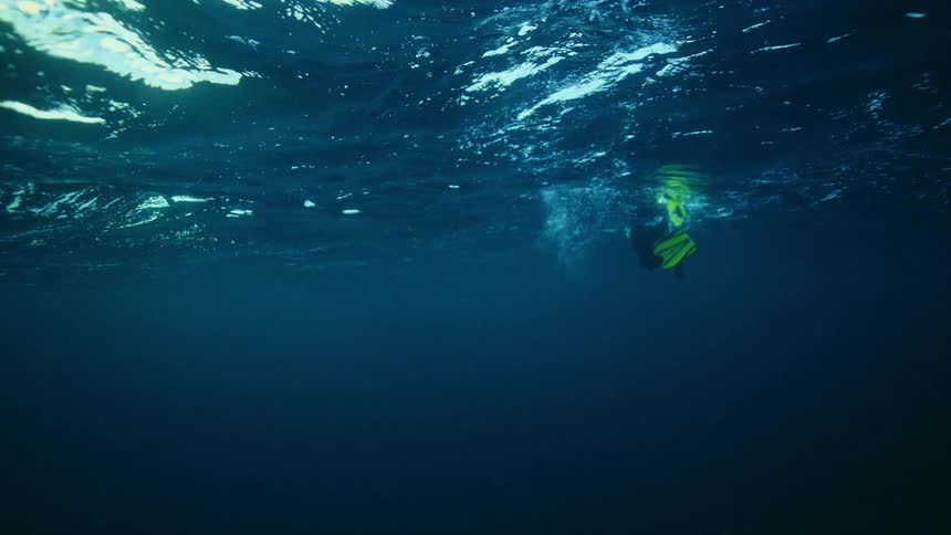 An underwater slow-motion view looking up at a scuba diver floating and kicking on the sunlit surface of the deep blue ocean