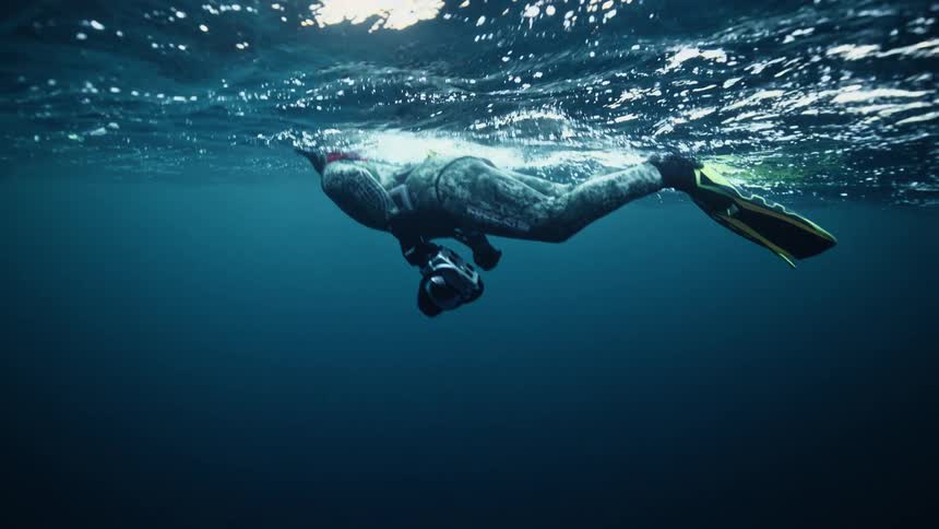 An underwater photographer in a wetsuit and flippers floats on the calm ocean surface in slow motion