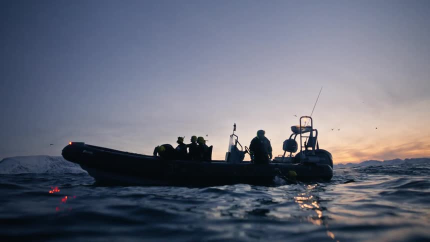 A silhouetted expedition boat with a crew floats on the calm arctic ocean, viewed from the water's surface at sunset