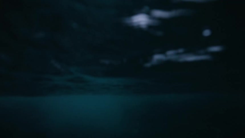 A slow-motion, split-level shot captures dark ocean waves lapping against a snow-covered mountain on an overcast day