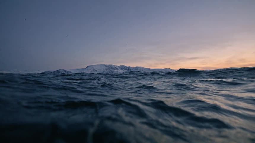 A serene, slow-motion view from the ocean surface reveals majestic snow-capped mountains at dusk