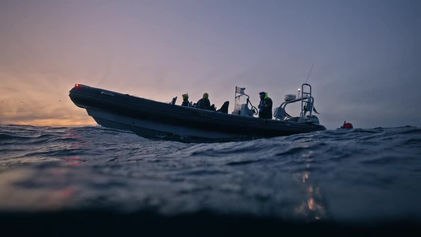 A slow-motion, half-submerged view reveals a boat of snorkelers rocking on the ocean waves against a sunrise sky and distant mountains