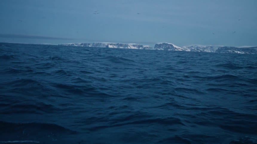 Seagulls fly over the vast, choppy blue sea as it rolls towards a distant, snow-covered coastline in slow motion