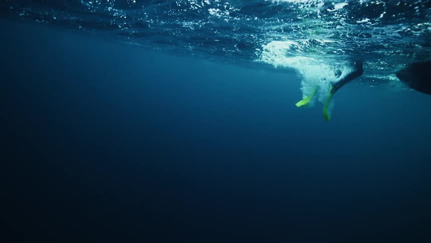 A slow-motion underwater view reveals a scuba diver descending into the deep blue ocean from the surface
