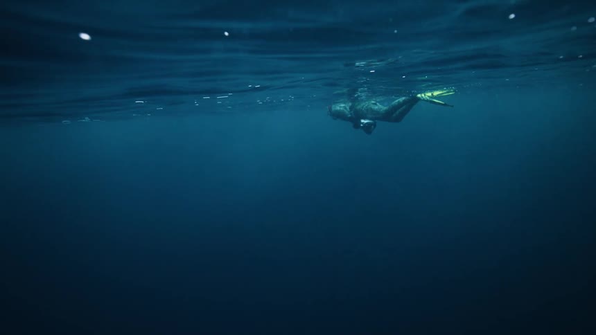 An underwater view shows a freediver wearing a wetsuit and fins floating on the surface of the vast, deep blue ocean