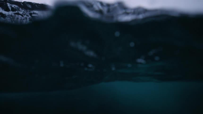 An atmospheric split-shot view from within dark, cold water reveals gentle waves and a snowy mountain shoreline