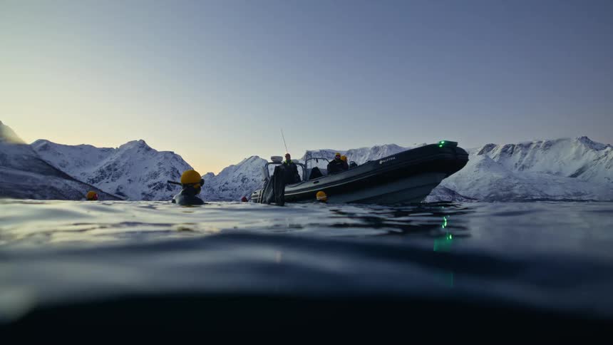 A split shot from the water's surface shows a dive boat and divers in an arctic fjord with snowy mountains in the background