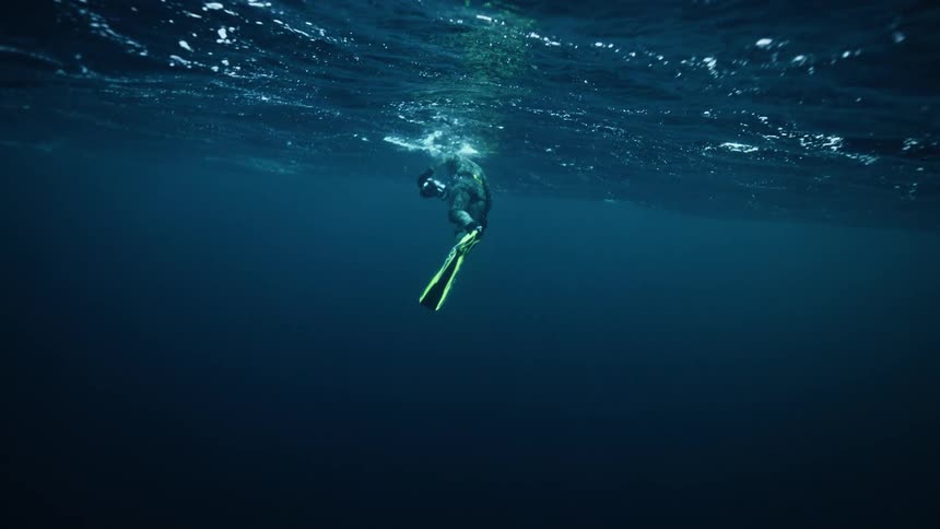 A lone freediver in a wetsuit and bright fins floats just beneath the ocean surface in slow motion