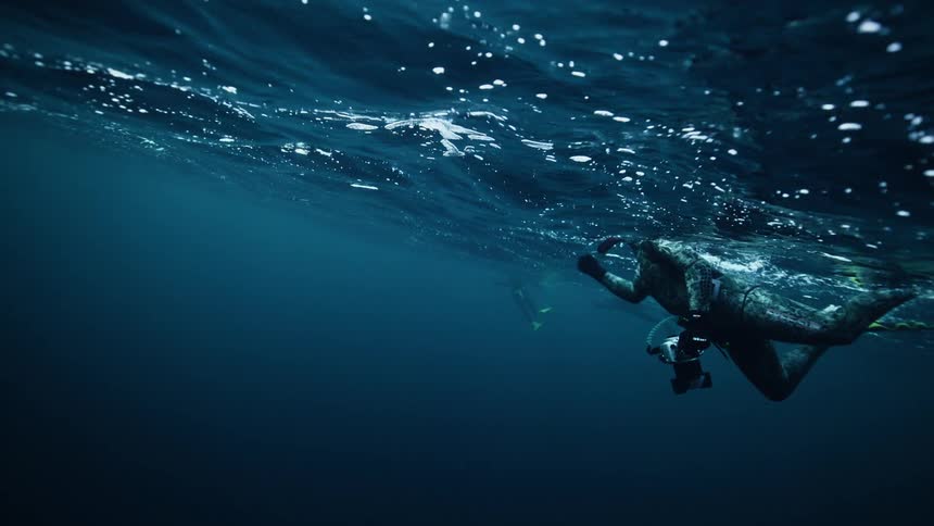 Slow-motion underwater view of a spearfisher in a full wetsuit floating peacefully just beneath the shimmering surface of the deep blue sea