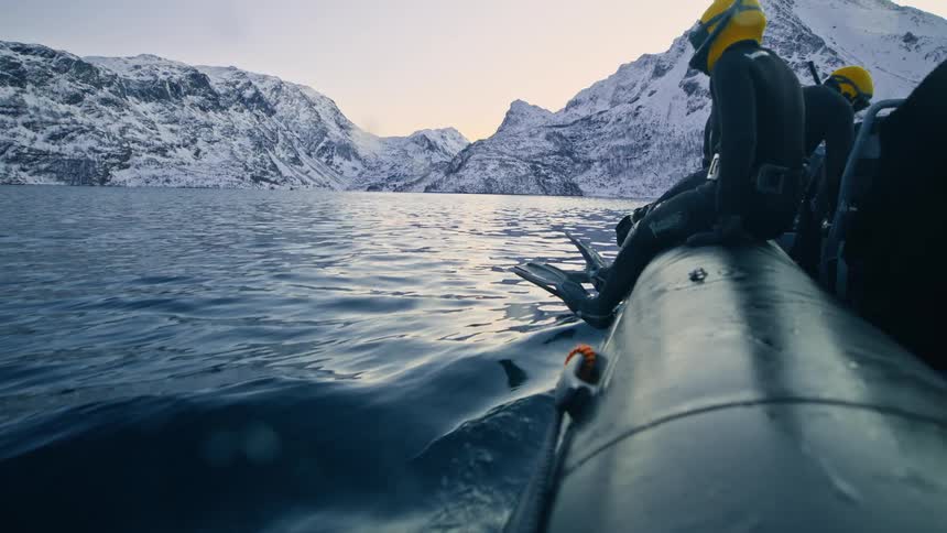 Divers in wetsuits ride an inflatable boat across a frigid arctic fjord surrounded by majestic snow-covered mountains