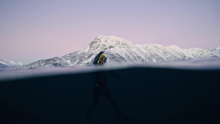 Cinematic split-level view of a swimmer in a wetsuit enjoying the frigid waters of a fjord with snowy mountains