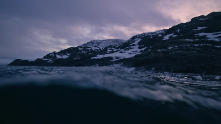 A moody split-level view shows dark ocean waves rolling in slow motion against a snowy shoreline under a dramatic sky