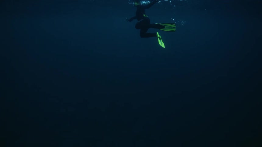 A lone scuba diver with bright yellow fins rests just below the surface in the vast, deep blue ocean
