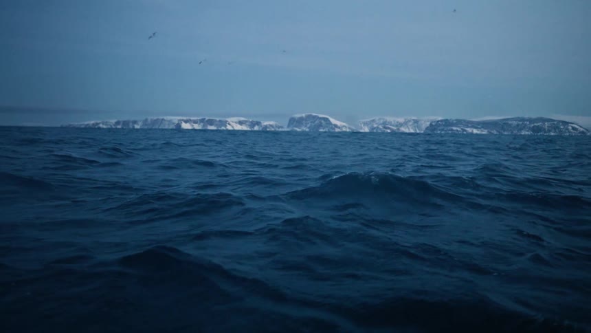 A slow-motion view from sea level captures the turbulent dark blue waters of the arctic ocean with a snowy coastline in the distance