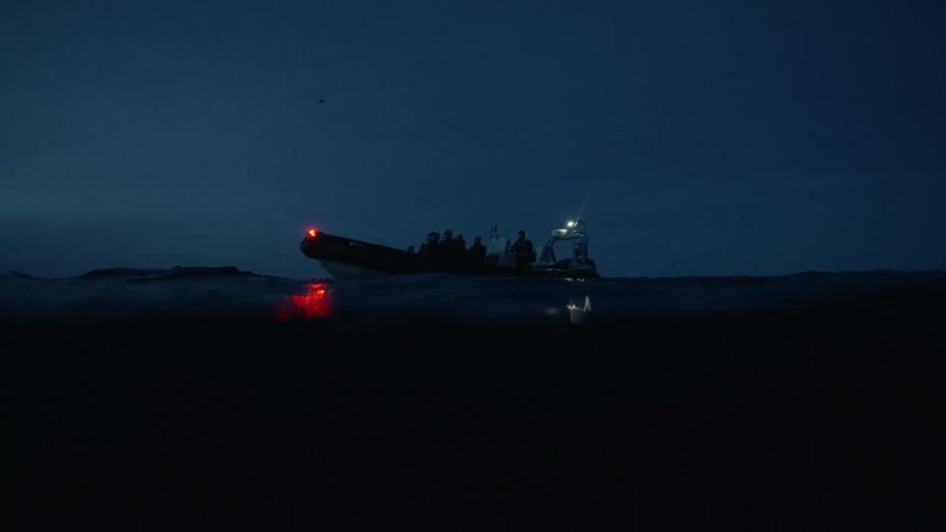 An immersive, slow-motion view from the surface of the dark ocean shows a tour boat floating at twilight
