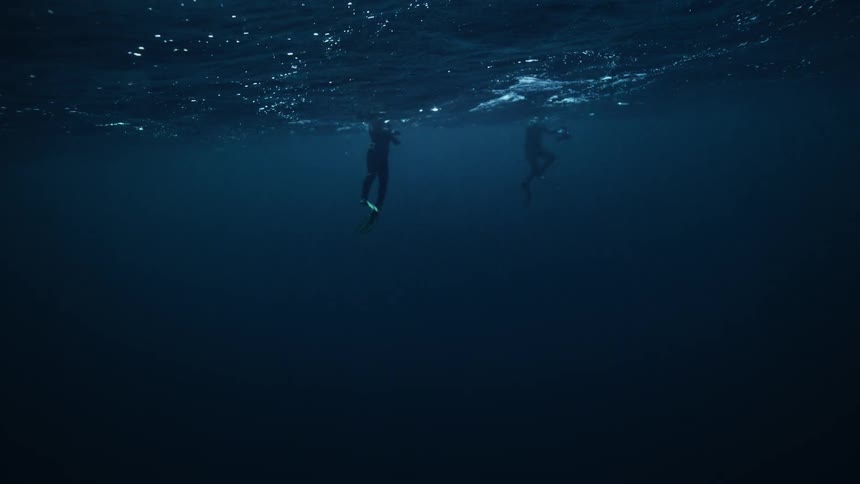 An underwater view from the dark depths of the ocean shows two divers floating on the rippling surface in slow motion