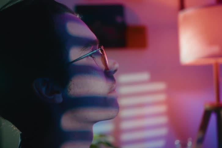 Close-up profile of a pensive young man in glasses with dramatic shadows from blinds cast across his face in a neon-lit room
