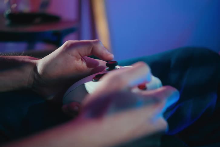 Close-up of a person's hands moving the joystick on a white video game controller in a dark room with blue and purple light