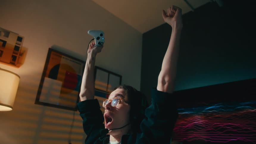 An ecstatic young gamer wearing a headset throws his hands up in victory while playing a video game at home