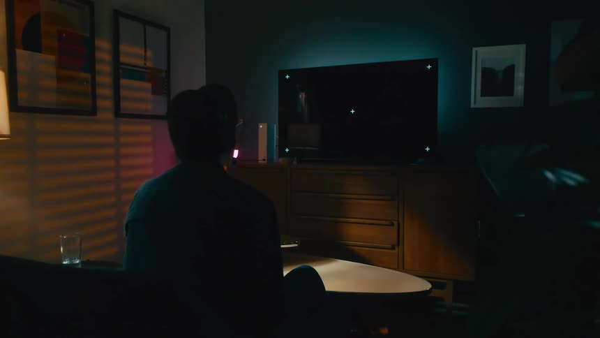 A gamer wearing headphones plays a video game on a large TV with a blank screen in a dimly lit, stylish living room