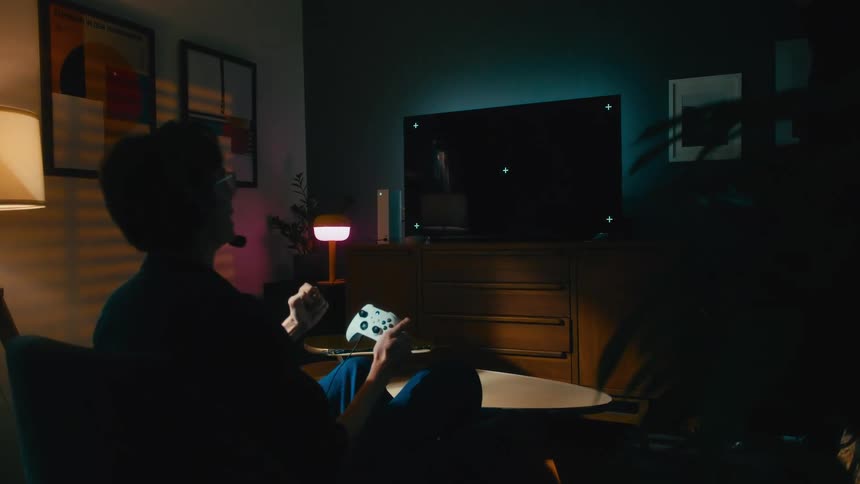An excited gamer wearing a headset celebrates winning while playing a video game on a console in his dark living room