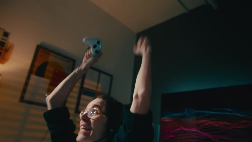 An excited young gamer wearing a headset and glasses celebrates a video game victory by triumphantly raising his arms and controller in the air in slow motion