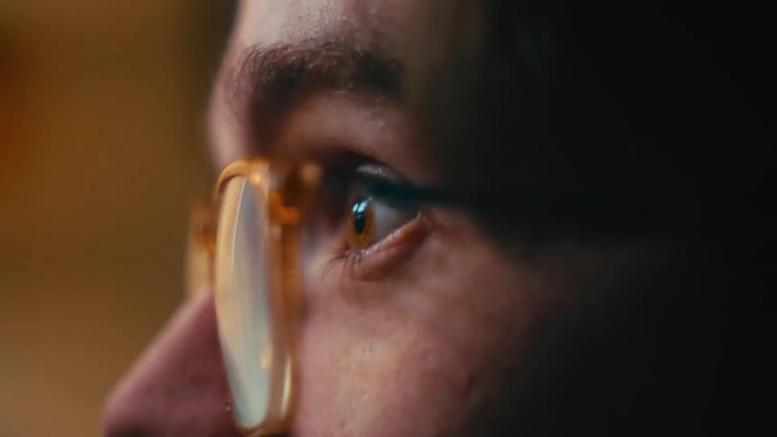 Intense macro shot of a person's eye, with a screen's glow reflecting in their glasses in a dark room