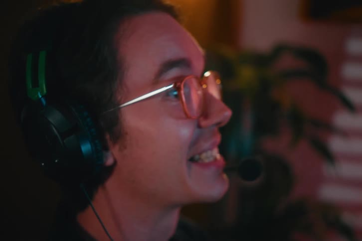 A close-up shot of a young man with glasses and a headset smiling and playing a video game at night