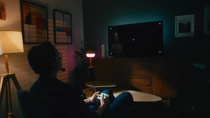 Gamer wearing a headset plays a video game with a controller in a dark living room in front of a blank television screen with tracking markers