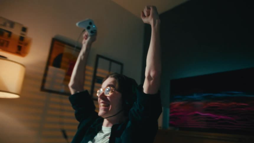 Triumphant young gamer with glasses and a headset celebrates winning his video game, raising his arms in the air