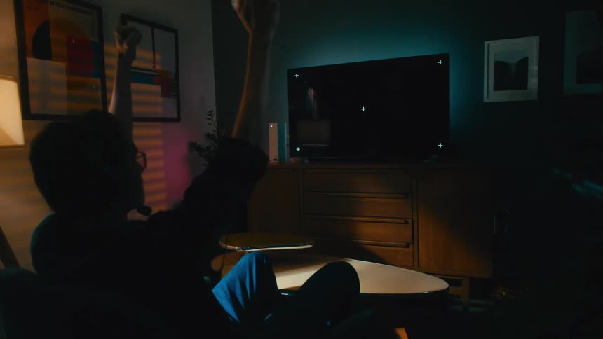 From behind, a young man plays a video game at night, celebrating a win with an excited fist pump in front of a TV with tracking markers