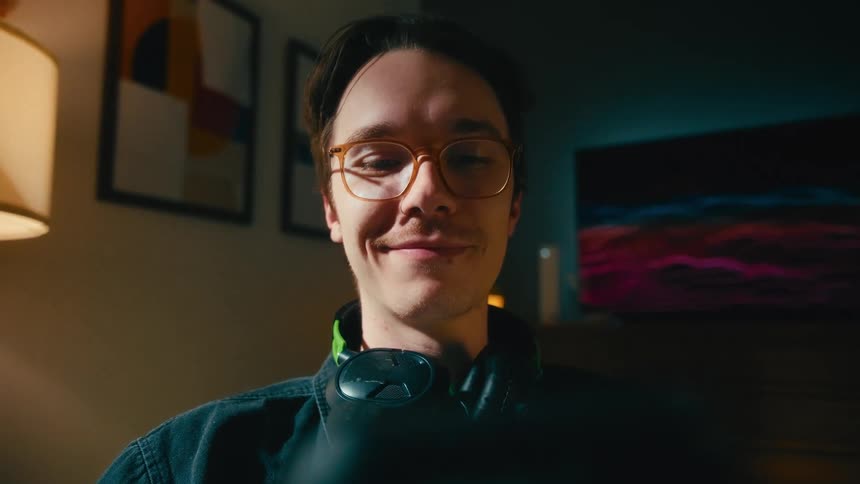 A young man with glasses looks up from his phone and smiles contentedly in a warmly lit room at night, POV